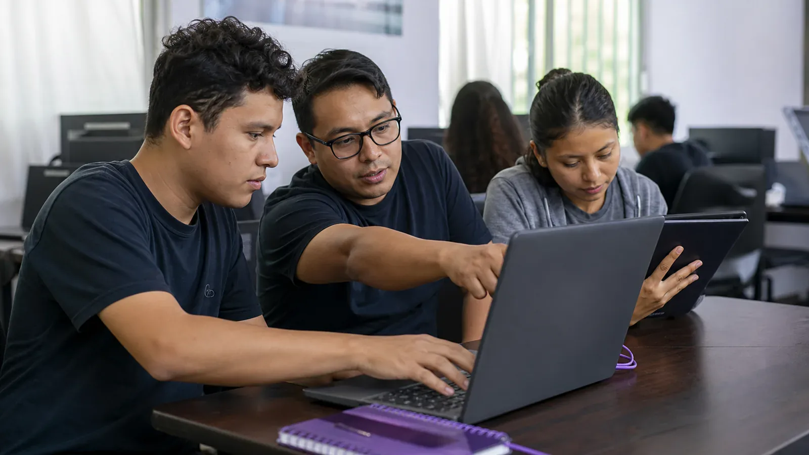 Un mentor y dos estudiantes trabajan juntos en un proyecto digital práctico con una laptop y una tableta.